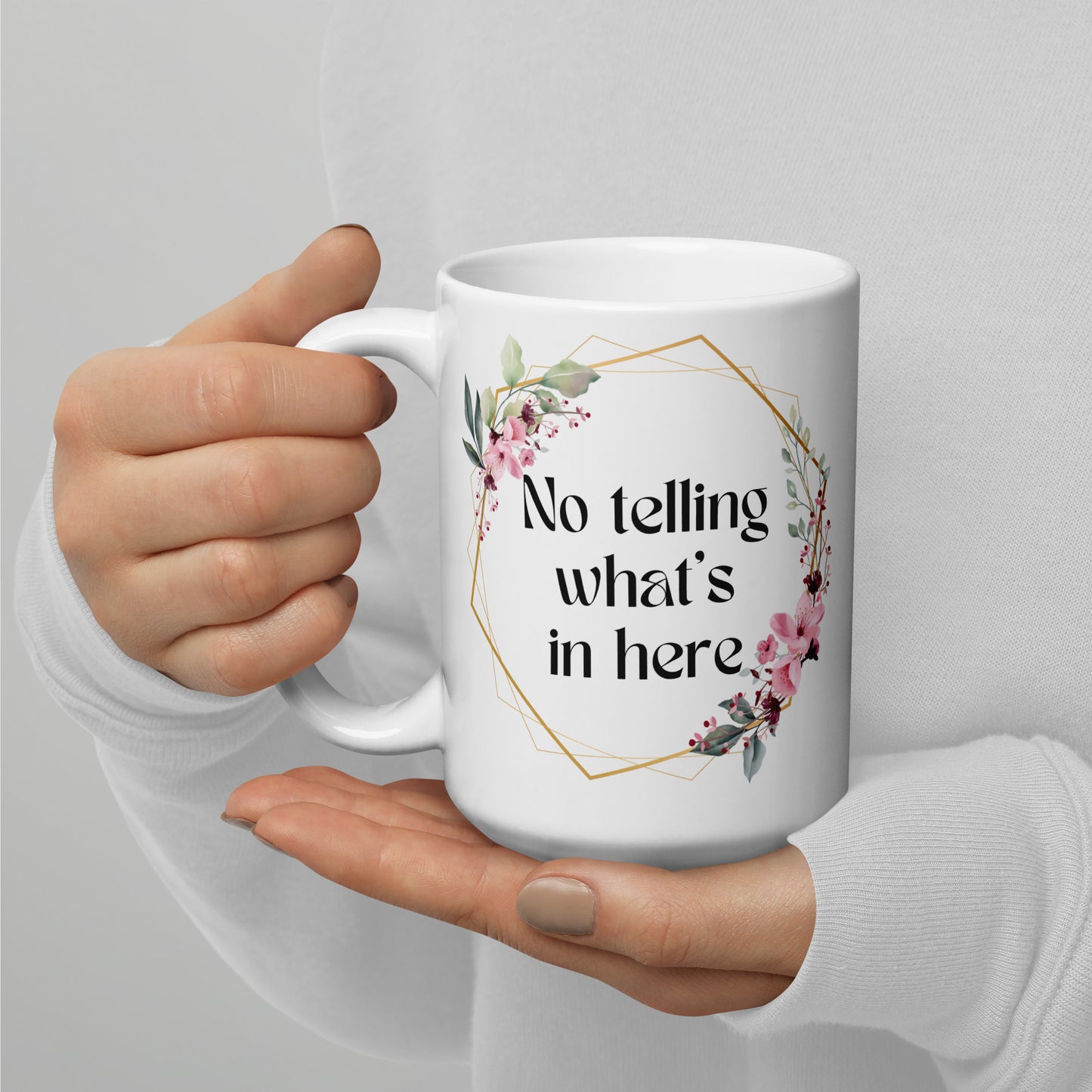 Manicured ands hold a white ceramic mug with a floral boho wreath that surrounds the text "NO TELLING WHAT'S IN HERE".