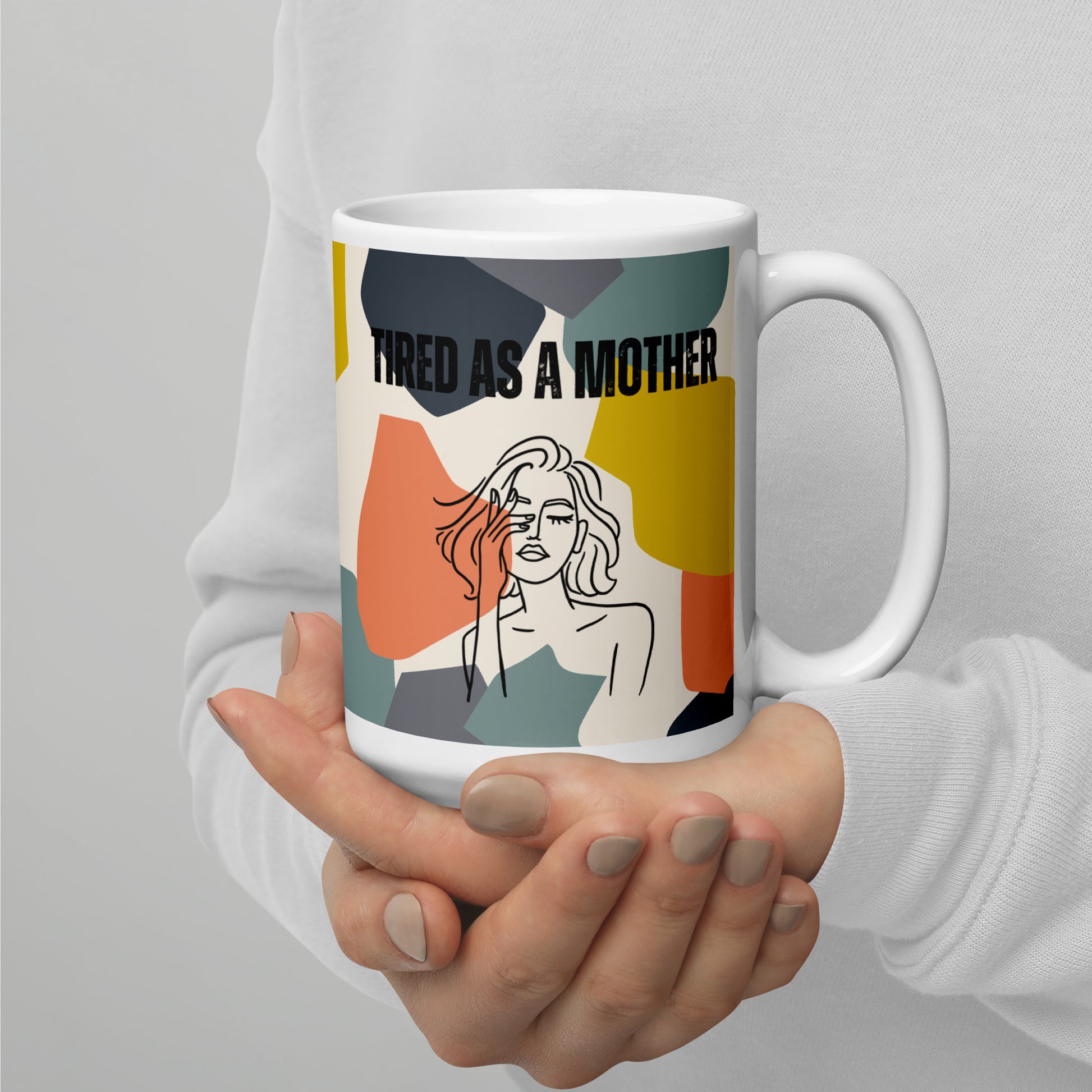 Manicured hands cup a mom's white ceramic mug - white ceramic mug featuring the text "TIRED AS A MOTHER" and an abstract illustration of a weary woman with tousled hair. The design incorporates a modern, colorful geometric background with shapes in yellow, orange, gray, and teal.