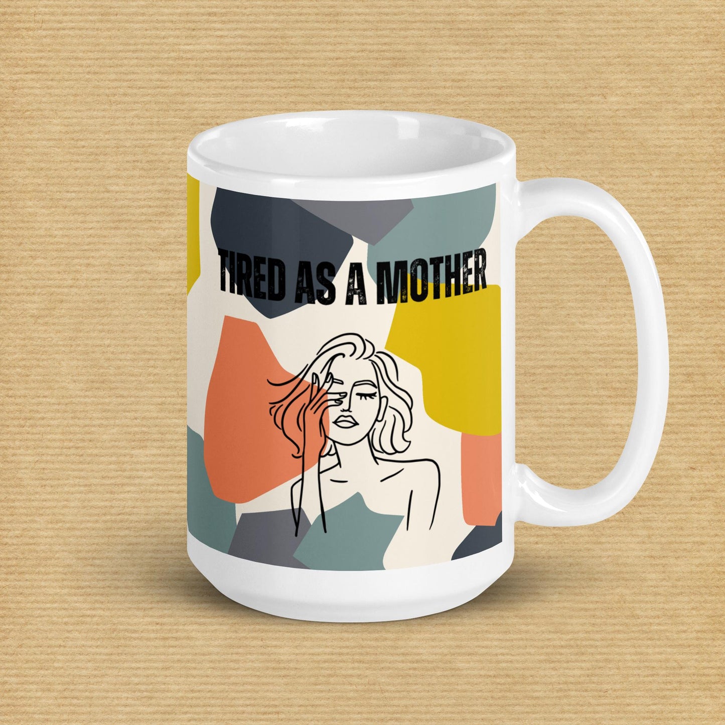 Colorful mug sits in front o a tan background - white ceramic mug featuring the text "TIRED AS A MOTHER" and an abstract illustration of a weary woman with tousled hair. The design incorporates a modern, colorful geometric background with shapes in yellow, orange, gray, and teal.