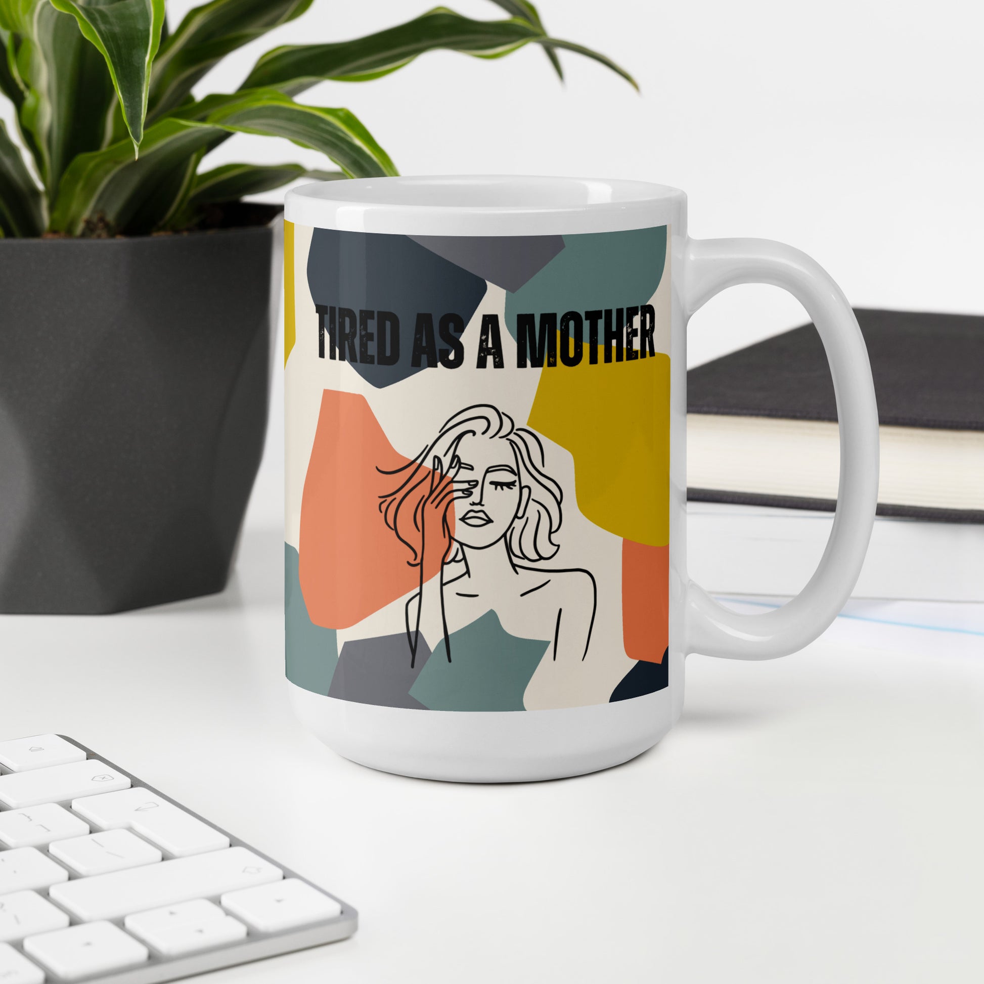 White ceramic mug with "TIRED AS A MOTHER" text and stylized illustration of a weary woman, set against a colorful geometric background. The mug is displayed on a desk next to a plant, keyboard, and book, showcasing its use in a home office setting.