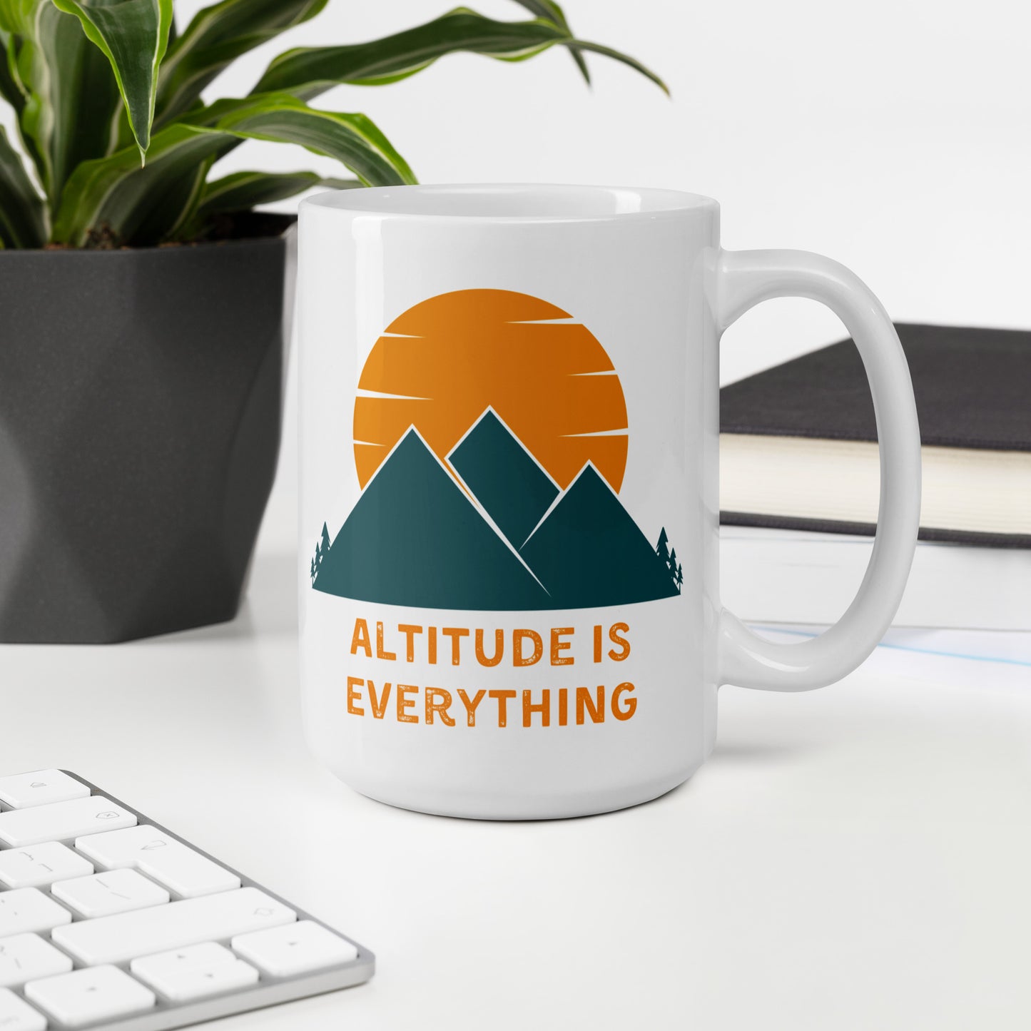 A 15 oz white ceramic mug with a graphic design of mountains and a sunset, along with the text "ALTITUDE IS EVERYTHING" sits on a desk next to a plant and a computer keyboard.