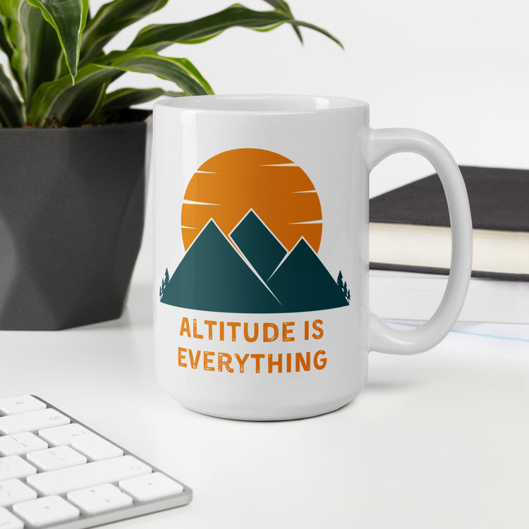 A 15 oz white ceramic mug with a graphic design of mountains and a sunset, along with the text "ALTITUDE IS EVERYTHING" sits on a desk next to a plant and a computer keyboard.
