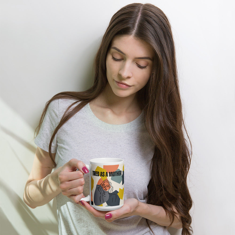 Youn woman with long brown hair and pink manicured hands cups a white ceramic mug with "TIRED AS A MOTHER" text and stylized illustration of a weary woman, set against a colorful geometric background. 