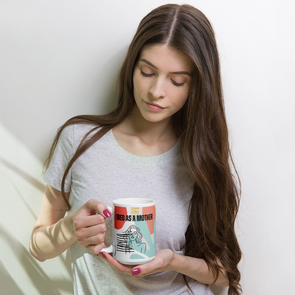 Woman with long brown hair and manicured fingernails holds a colorful coffee mug that reads "TIRED AS A MOTHER". Under the text is line art of a woman with her eyes closed, rubbing one eye. A colorful background pattern includes mint, rosy pink, yellow, coral and black with shapes reminiscent of the 80s or early 90s.