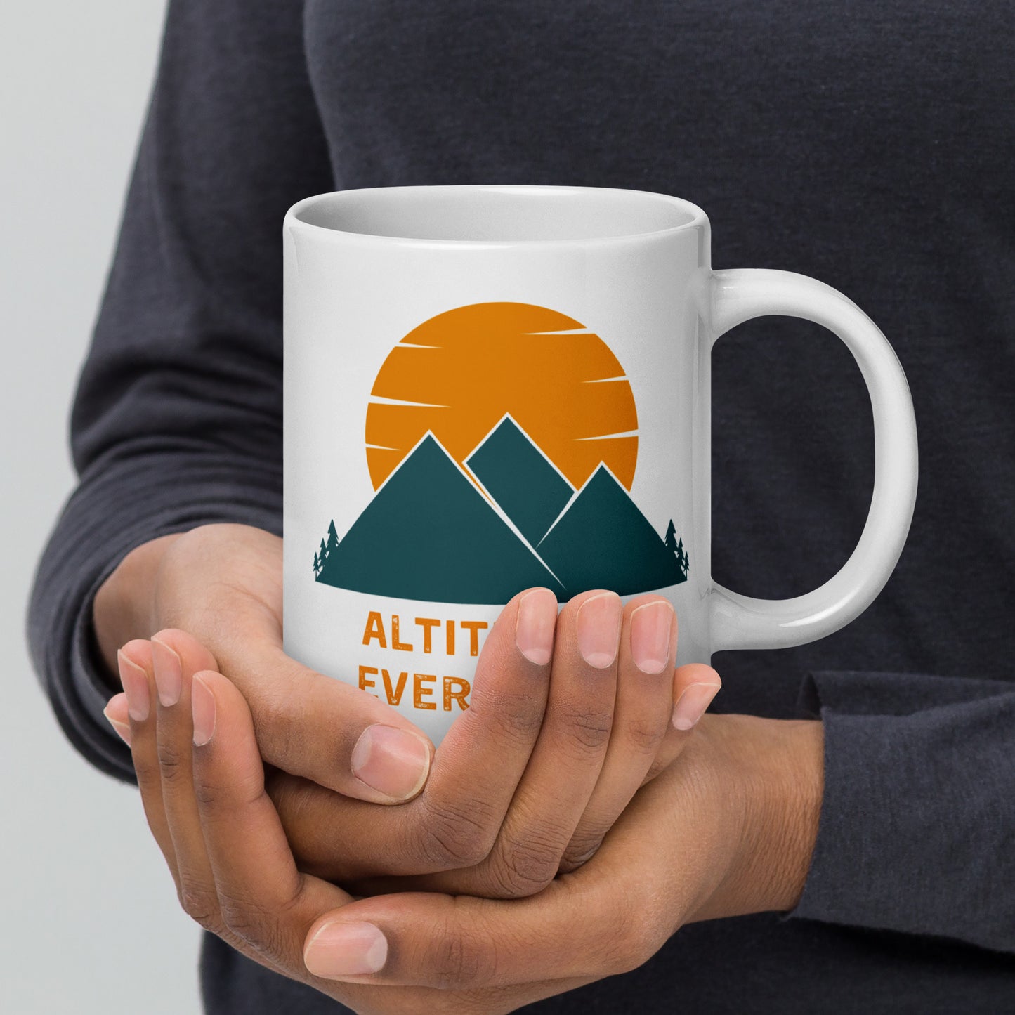 A person in a dark gray shirtt cups a white ceramic mug with mountains and a sunset, along with the text "ALTITUDE IS EVERYTHING",