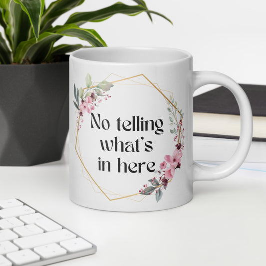 A white ceramic mug with a floral wreath design and the text "No telling what's in here" displayed prominently. The mug is placed on a desk next to a computer keyboard, with a potted plant in the background, creating a cozy, office-like setting. The mug's design features a delicate floral arrangement in soft pink and green hues, giving it a bohemian, feminine aesthetic.