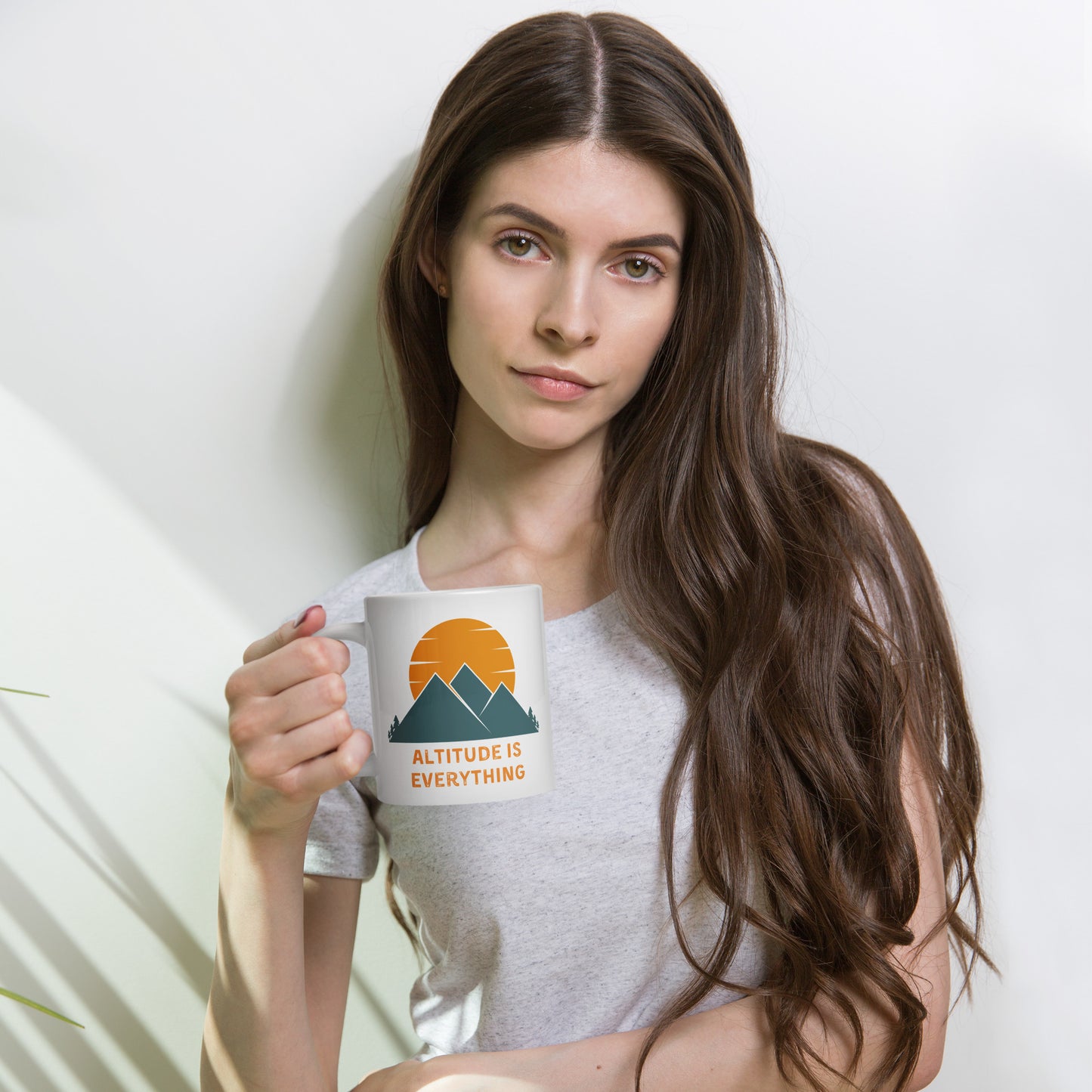 Woman with ling brown hair holds a white ceramic mug that reads Altitude is Everything. The mug also displays a graphic of mounatins and a sunset with orange and deep blue coloring.