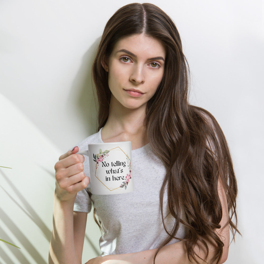 Woman with long brown hair hold a  white ceramic mug with a floral wreath design and the text "No telling what's in here". The mug's design features a delicate floral arrangement in soft pink and green hues, giving it a bohemian, feminine aesthetic.