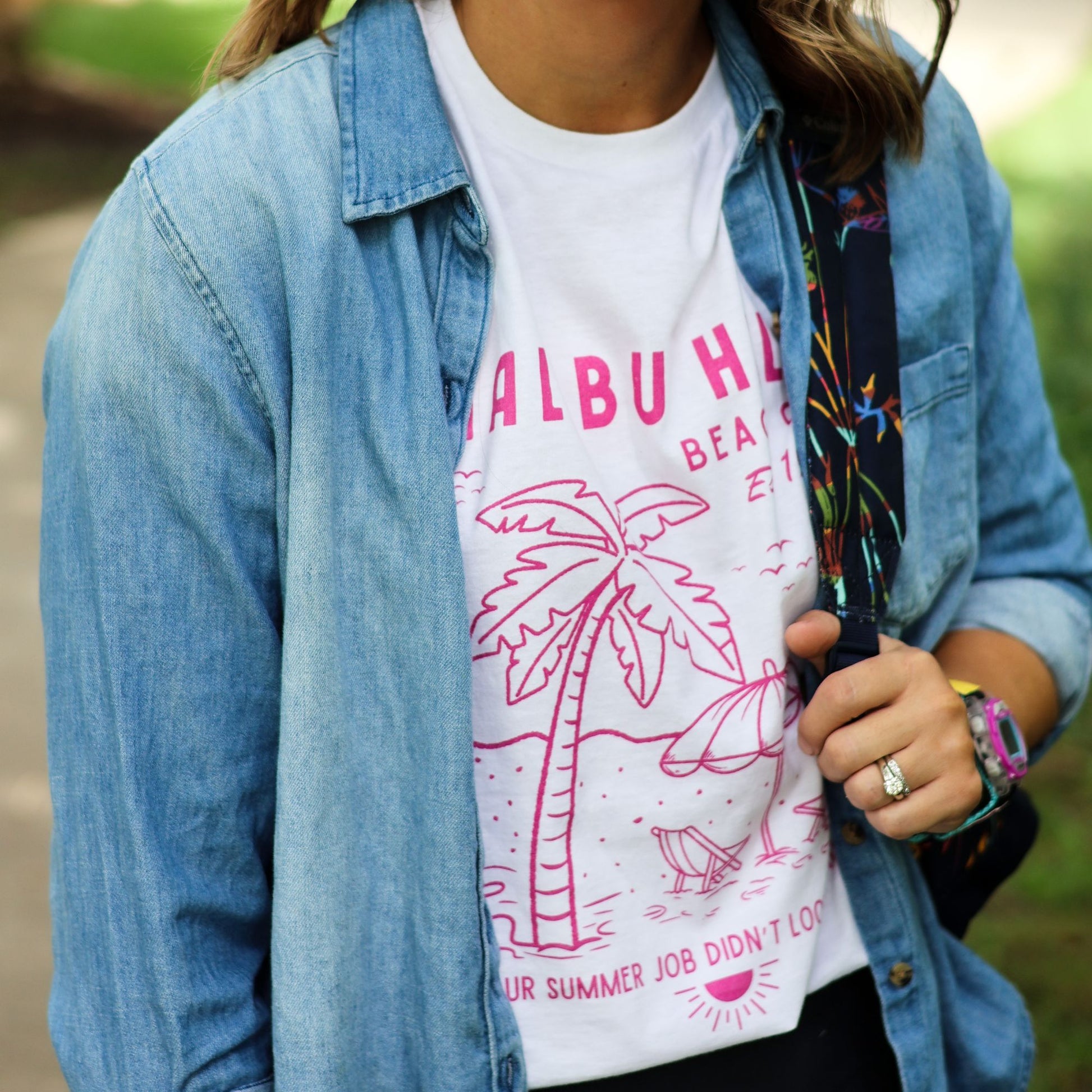 Woman outdoors wearing a white t-shirt with pink graphics and text, paired with a denim shirt and shoulder bag.