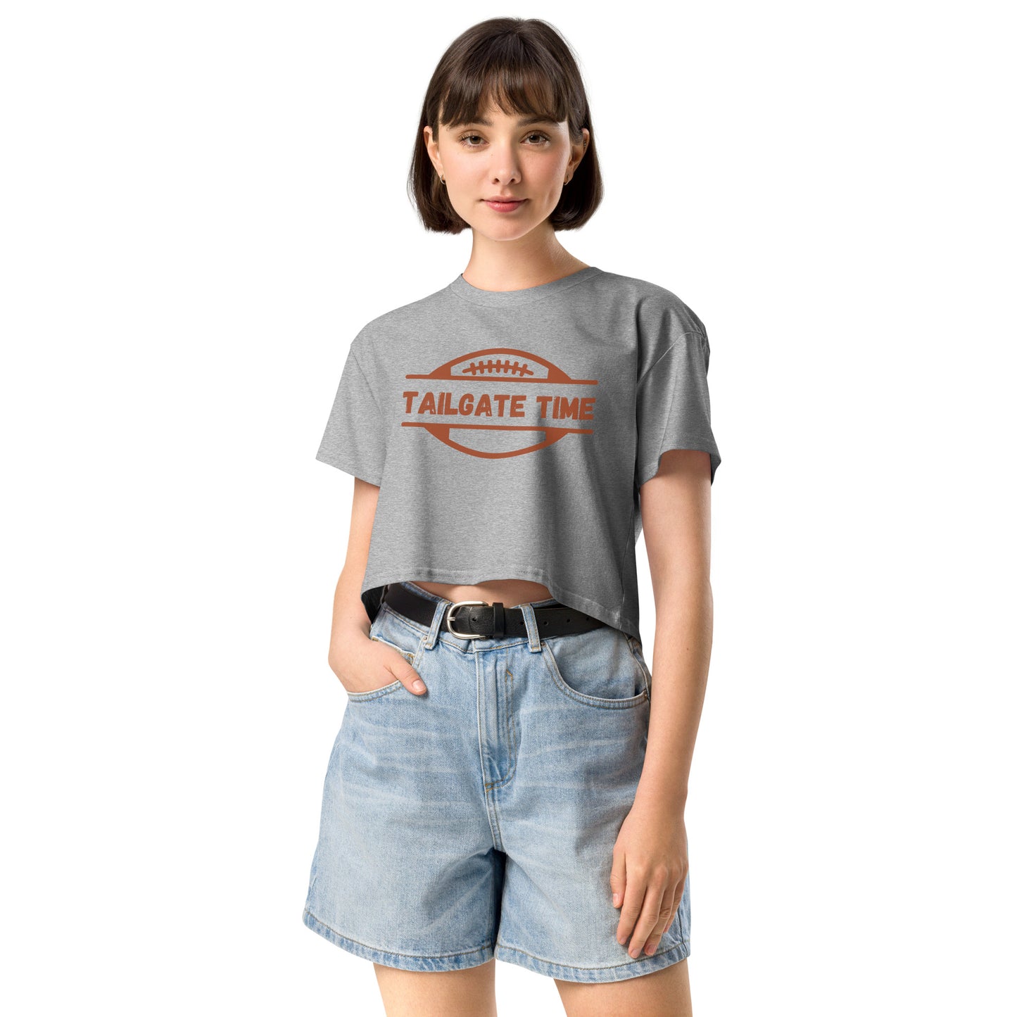 Young woman wears a football crop top and high waisted jean shorts. The crop top has a football with words "Tailgate Time" in distressed lettering.