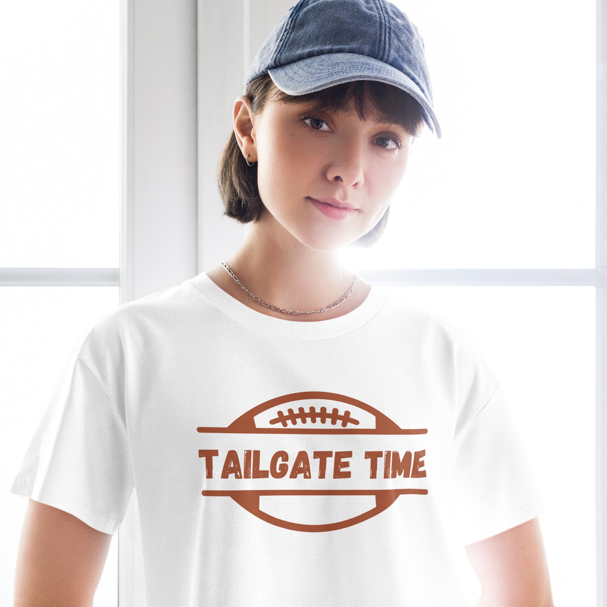 Young woman wears a football crop top, necklace and a denim ball cap. The crop top has a football with words "Tailgate Time" in distressed lettering.