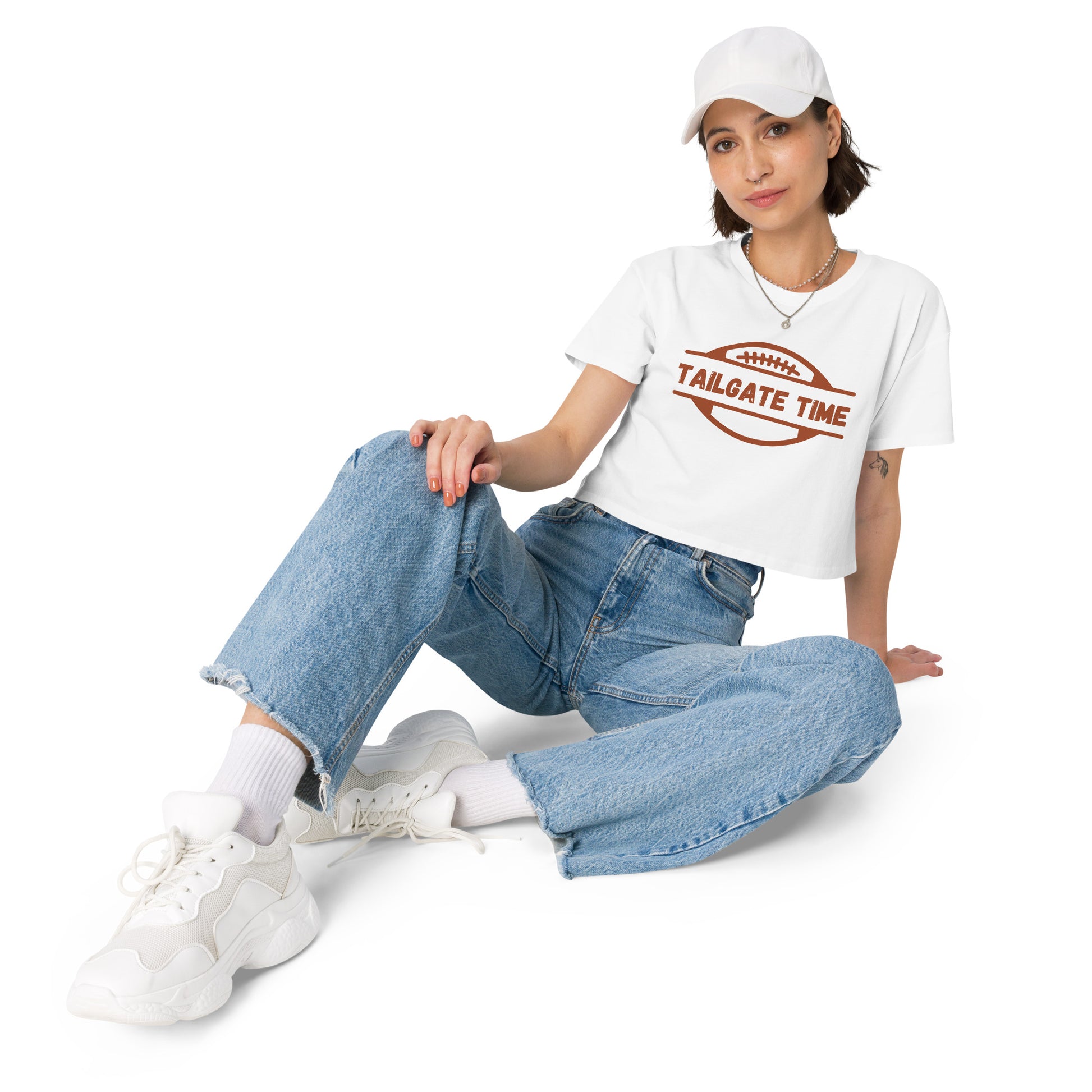 Young woman wears a football crop top, high waist jeans, white sneakers, and a while ball cap. The crop top has a football with words "Tailgate Time" in distressed lettering.