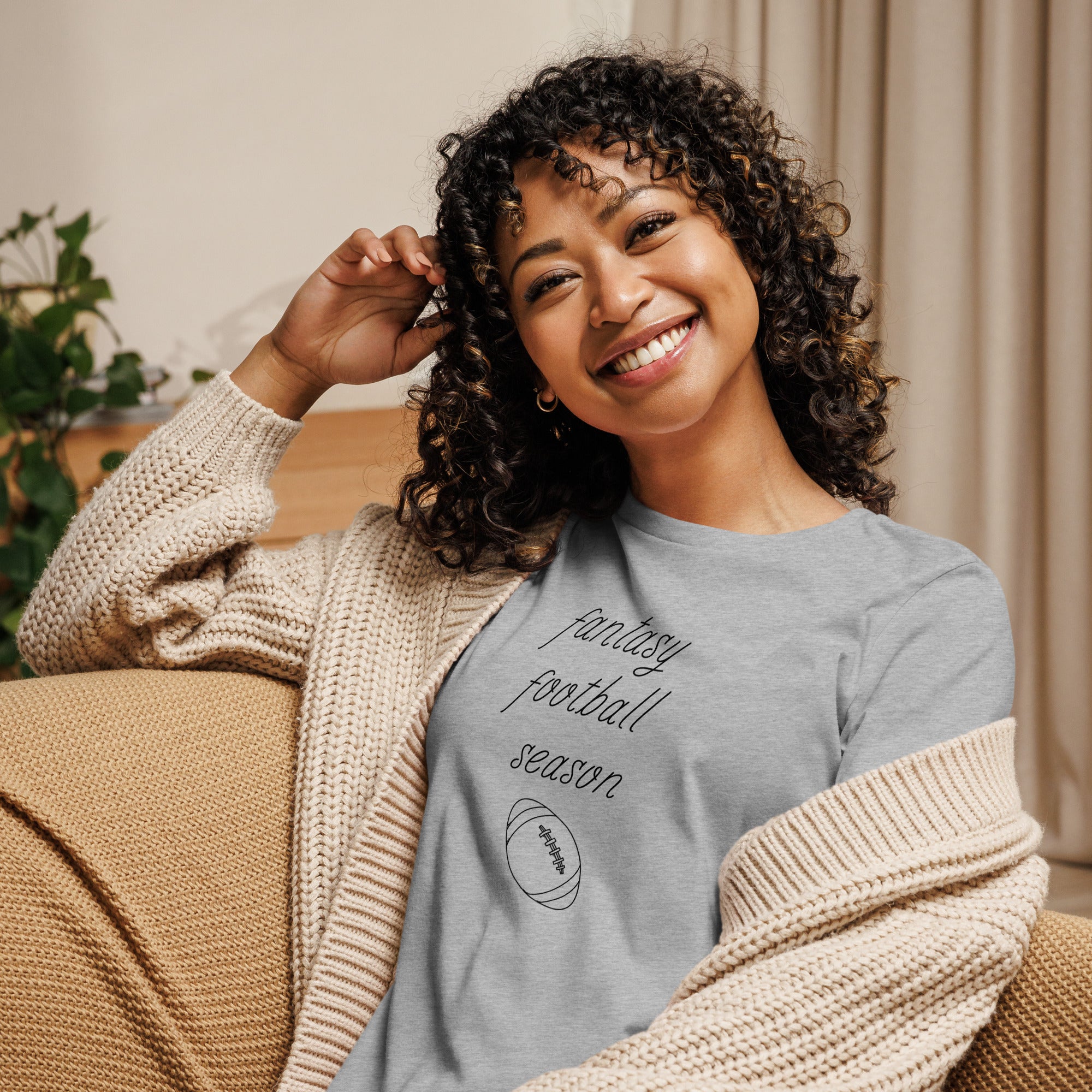 Smiling woman with curly hair sits on a couch wearing a tee  shirt that reads "FANTASY FOOTBALL SEASON" along with an image of a football. The font is a thin and flowy cursive that is pretty and feminine.