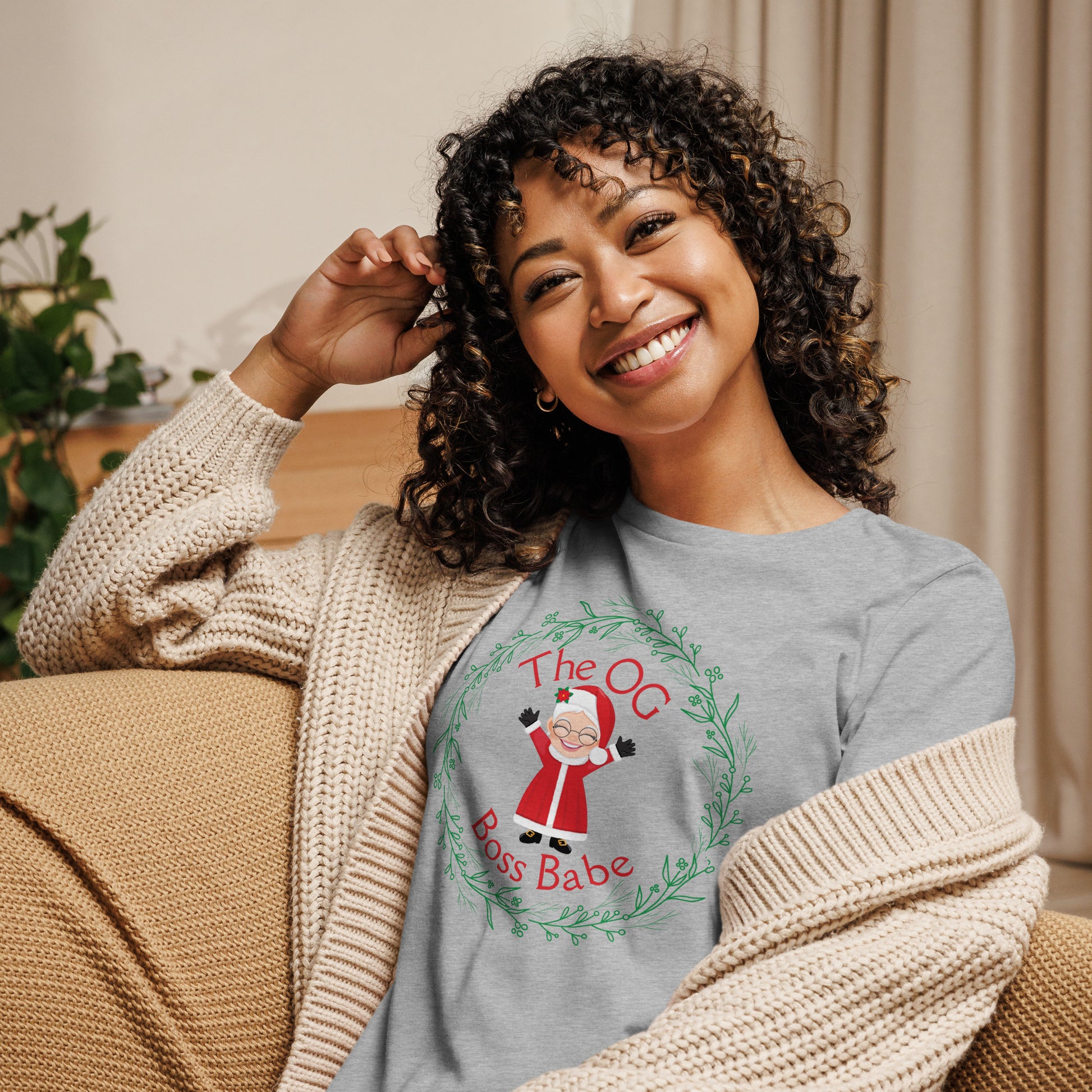 A smiling woman wears a t-shirt that features a graphic of Mrs. Claus with the text "The OG Boss Babe" printed around her. The empowering t-shirt has a festive, holiday-themed design that suggests Mrs. Claus is the true force behind the magic of Christmas.