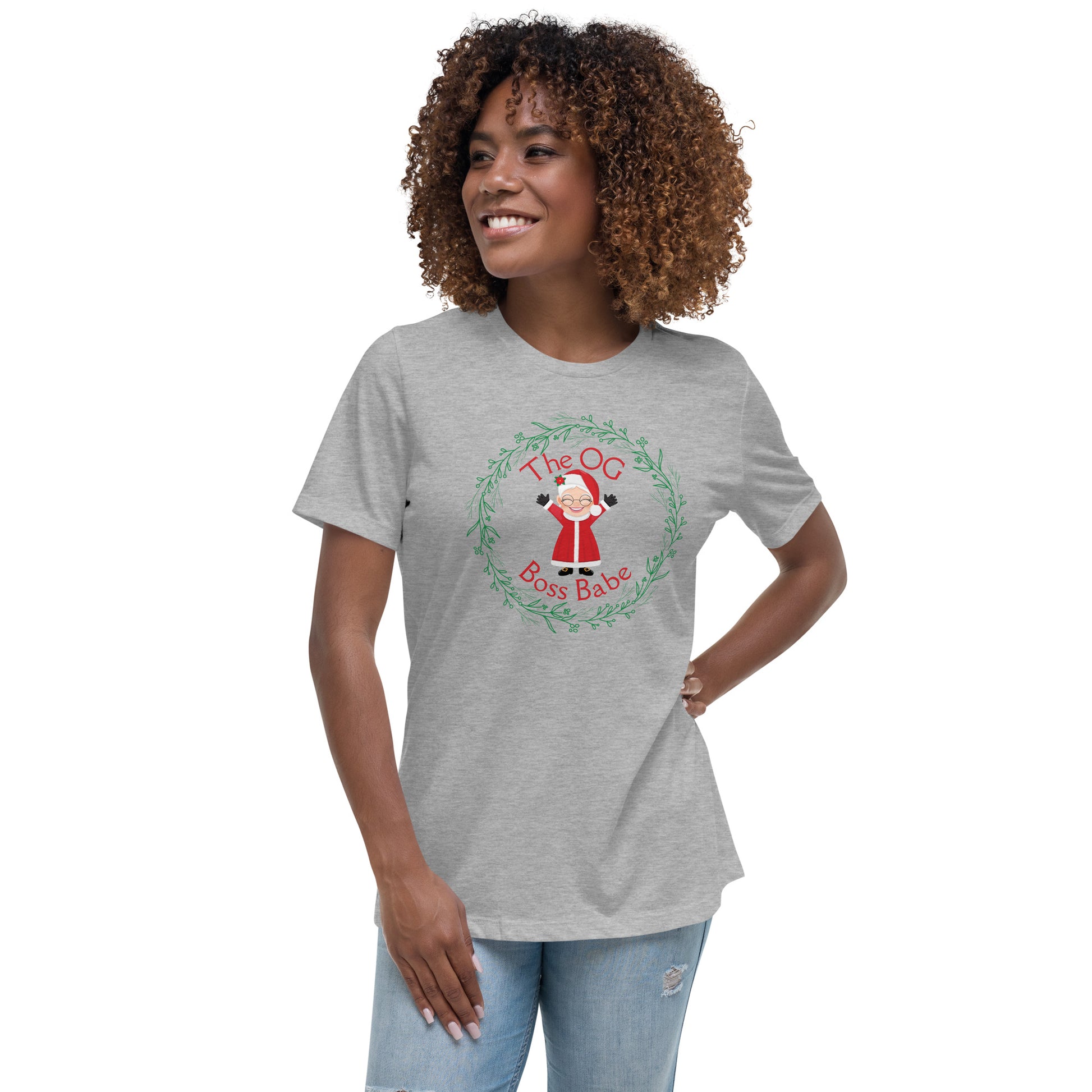 A smiling woman wears a t-shirt that features a graphic of Mrs. Claus with the text "The OG Boss Babe" printed around her. The empowering t-shirt has a festive, holiday-themed design that suggests Mrs. Claus is the true force behind the magic of Christmas.