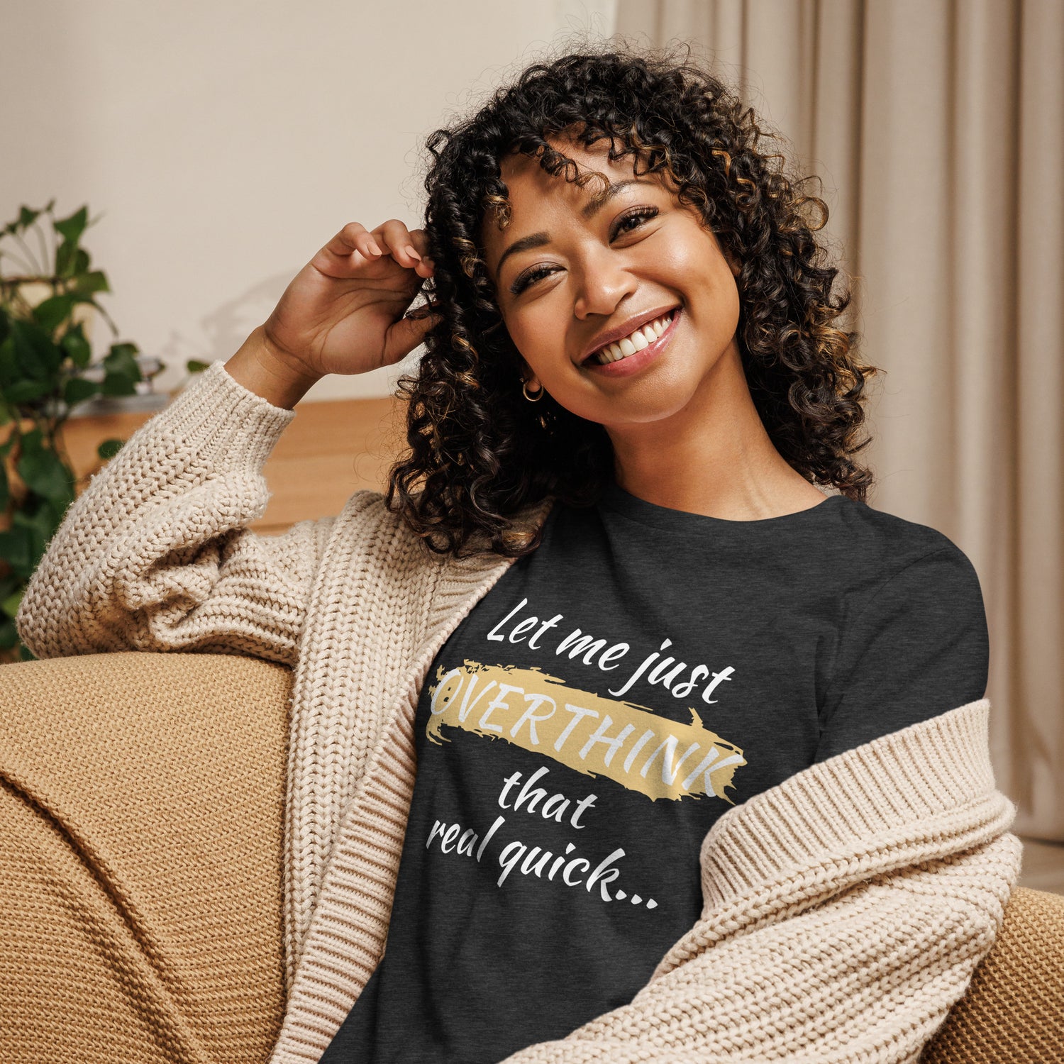 Smiling young woman with curly hair sits on a couch wearing a graphic tee shirt that reads "Let me just OVERTHINK that real quick..."