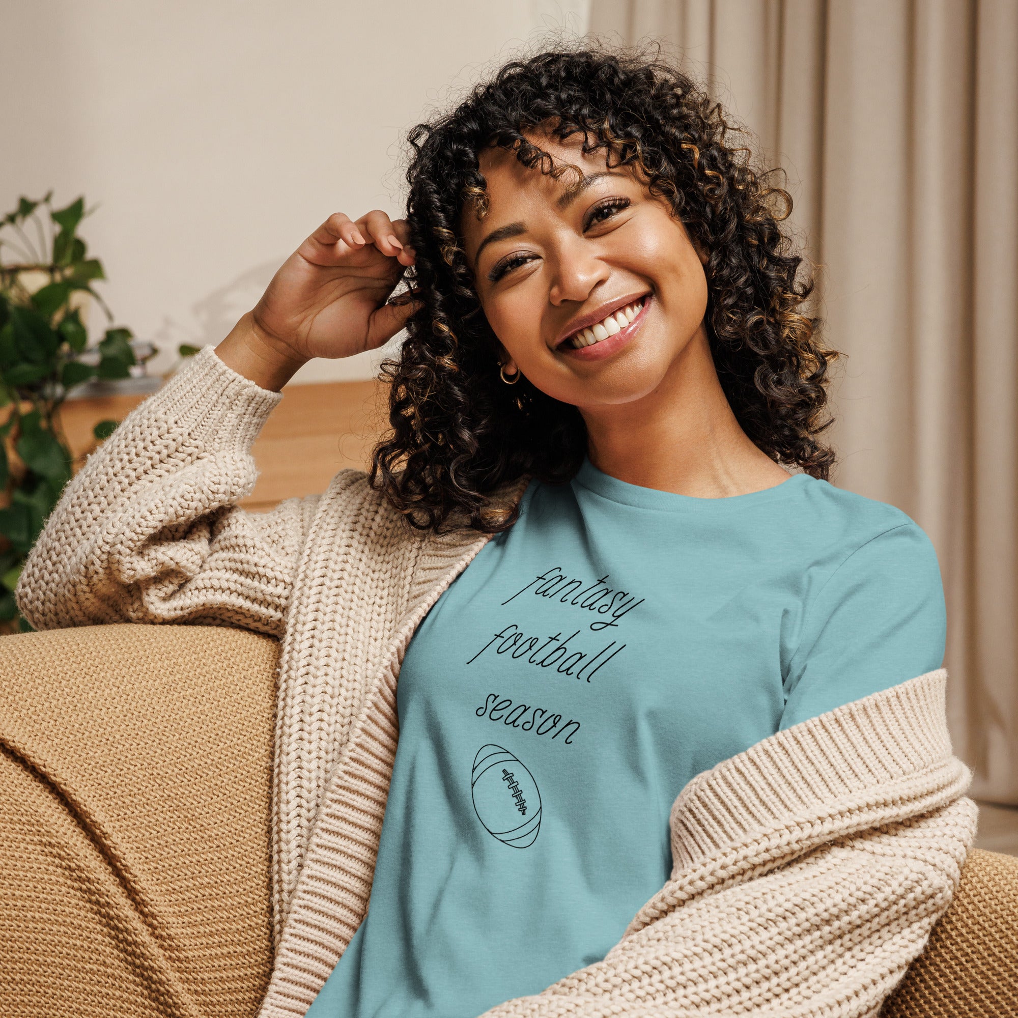 Smiling woman with curly hair sits on a couch wearing a tee  shirt that reads "FANTASY FOOTBALL SEASON" along with an image of a football. The font is a thin and flowy cursive that is pretty and feminine.