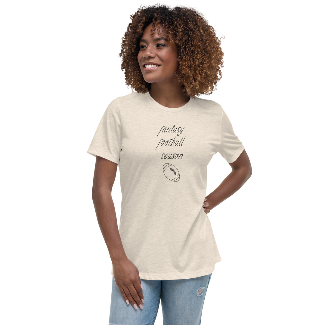 Woman in jeans sports an off-white women's tee shirt that reads "FANTASY FOOTBALL SEASON" along with an image of a football. The font is a thin and flowy cursive that is pretty and feminine.