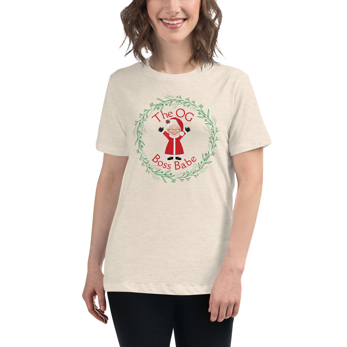 A smiling woman wears a t-shirt that features a graphic of Mrs. Claus with the text "The OG Boss Babe" printed around her. The empowering t-shirt has a festive, holiday-themed design that suggests Mrs. Claus is the true force behind the magic of Christmas.