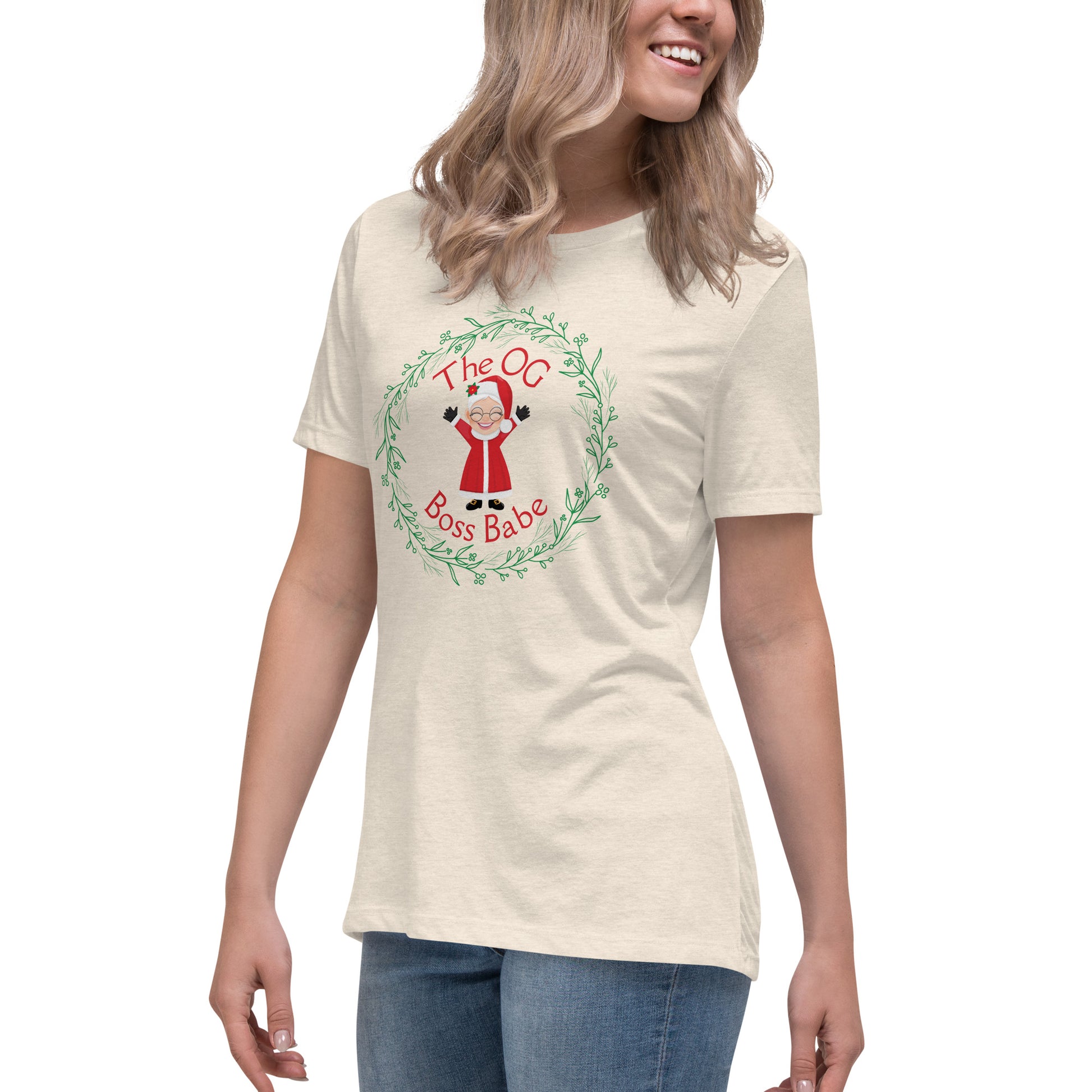 A smiling woman wears a t-shirt that features a graphic of Mrs. Claus with the text "The OG Boss Babe" printed around her. The empowering t-shirt has a festive, holiday-themed design that suggests Mrs. Claus is the true force behind the magic of Christmas.